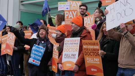 In-slow-motion-Junior-Doctors-wearing-orange-hats,-hold-placards,-wave-blue-flags-and-cheer-on-a-picket-line-as-passing-vehicles-honk-horns-in-support-outside-the-University-College-London-Hospital