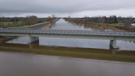 Aerial-establishing-view-of-high-water-in-springtime,-Barta-river-flood,-brown-and-muddy-water,-overcast-day,-wide-drone-shot-moving-forward-over-the-bridge