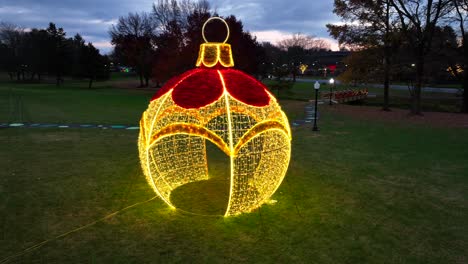 Aerial-orbiting-shot-of-large-lighting-Christmas-ornament-and-bulb-on-grass-field-in-American-park-during-dusk-in-December