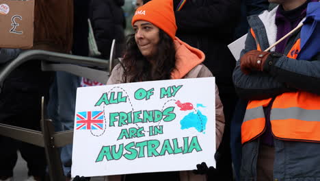 In-slow-motion-a-woman-holds-a-cardboard-placard-that-reads,-“All-of-my-friends-are-in-Australia”-on-a-strike-action-picket-line-outside-the-entrance-of-the-University-College-London-Hospital