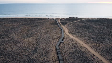 An-aerial-perspective-captures-the-cinematic-beauty-of-Portugal's-coastal-landscape,-featuring-a-walking-pathway-and-a-solitary-road-winding-through-a-desolate-terrain-to-water-surface