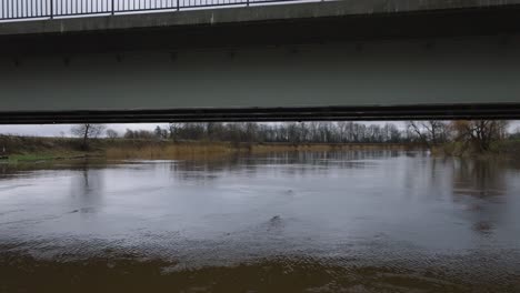 Aerial-establishing-view-of-high-water-in-springtime,-Barta-river-flood,-brown-and-muddy-water,-overcast-day,-low-drone-shot-moving-forward-below-the-concrete-bridge