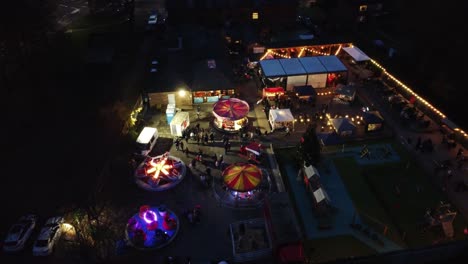 Illuminated-Christmas-fairground-festival-in-neighbourhood-car-park-at-night-aerial-orbit-view