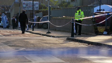 A-Metropolitan-police-officer-stands-guard-at-the-entrance-to-a-property-behind-a-gun-crime-murder-scene-cordon-as-two-forensic-officers-enter-in-Hackney