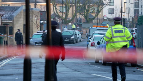 Metropolitan-police-officers-walk-through-a-gun-crime-murder-scene-cordon-as-others-stand-guard-at-the-entrance-to-a-property-in-Hackney