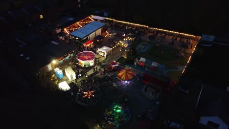 Illuminated-Christmas-fairground-festival-in-neighbourhood-car-park-at-night-aerial-circling-view