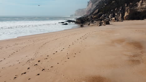 Flying-Above-Beach-Sand-With-Footprints-On-Rocky-Ocean-Coast-Of-Nazare-In-Leiria,-Portugal