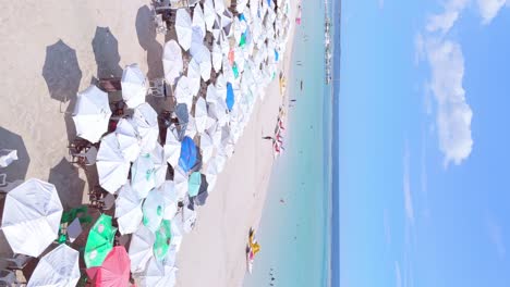 Portrait-aerial,-tourists-enjoying-tropical-summer-under-parasols,-sand-beach