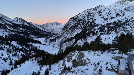Drone-footage-of-a-mountain-refuge-under-the-snow-at-dawn,-revealing-a-snowy-valley-in-the-Pyrenees-mountains