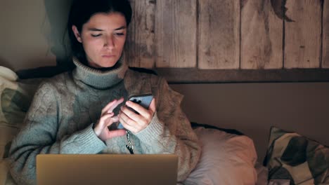 Young-Caucasian-woman-browsing-on-phone-and-laptop-while-lying-in-bed-at-home