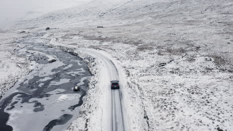 Färöer-Inseln,-4k-Luftverfolgung-Hinter-Einem-Schwarzen-Auto-Im-Schnee-Mit-Wunderschönen-Bergen-Im-Hintergrund