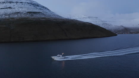Faroe-Islands,-4K-Aerial-tracking-pan-of-fishing-boat-with-snow-covered-mountains-in-the-background-as-it-dissapears-into-the-sun