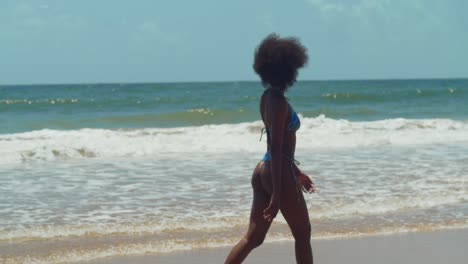 In-the-midst-of-a-tropical-paradise,-a-girl-with-curly-hair-walking-on-the-shoreline-on-a-Caribbean-island-beach-in-a-bikini-with-ocean-waves-in-the-background