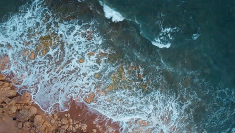 Drone-top-down-shot-of-waves-crushing-on-rocky-beach,-drone-moving-towards-the-water