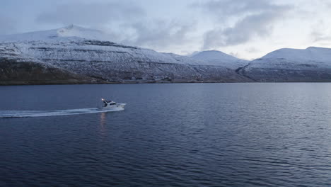 Faroe-Islands,-4K-Aerial-tracking-three-quaeter-shot-of-fishing-boat-with-snow-covered-mountains-in-the-background