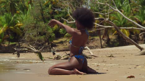 On-a-sunny-day,-a-girl-with-curly-hair-in-a-bikini-finds-joy-on-a-tropical-Caribbean-island-beach-kneel-in-the-sand