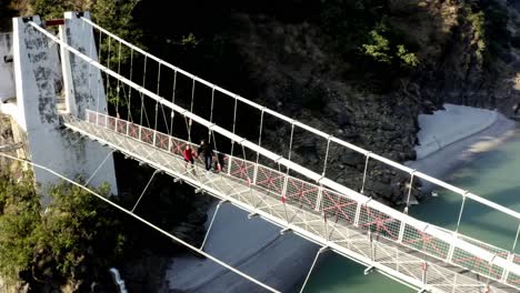 aerial-drone-camera-cinematic-shot-chopta-utrakhand-a-couple-is-walking-on-a-bridge-between-the-surrounding-mountains-with-water-below-it