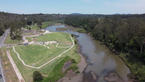 Drone-flying-over-a-highway-towards-a-green-recreational-park-with-a-small-brown-river-near-the-park