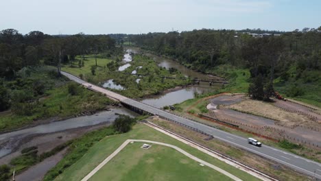 Drone-descending-on-to-a-green-park-showing-a-small-river-and-a-bridge-with-cars-passing-by