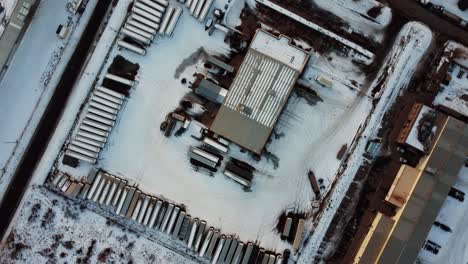 Top-down-aerial-view-of-trucks,-trailers,-and-tanks-parked-in-an-outdoor-warehouse-during-winter