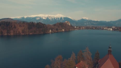 Drone-forward-flying-view-of-Bled-lake-island-and-church,-snow-covered-alps-in-distance