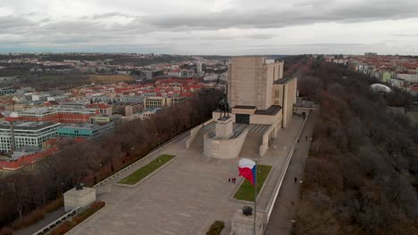 Vista-Aérea-Cinematográfica-Del-Punto-De-Referencia-De-Praga,-Monumento-Nacional-En-La-Colina-Vitkov-Con-La-Bandera-Checa-Ondeando-En-Una-Tarde-Ventosa