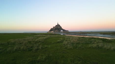 Aerial-Drone-Shot-of-Mont-Saint-Michel-Ascending-over-Grass-at-Beautiful-Orange-Sunset-in-4K