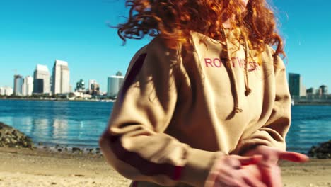 Urban-street-dancer,-red-hair-female-dancing-on-waterfront-with-views-in-background-of-high-rise-city-skyline