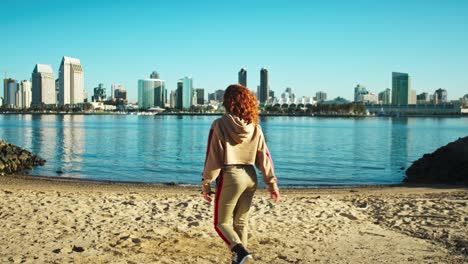 Long-red-haired-female-urban-dancer-walking-into-city-skyline-waterfront-scene-starting-to-dance-slow-motion-on-bright-sunny-blue-sky-day
