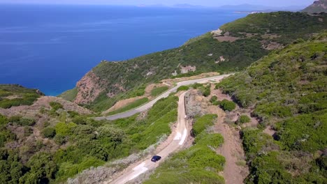 Drone-Aerial-shot,-following-a-car-driving-on-a-coastal-road-on-a-mediterranean-landscape-with-stunning-blue-water-and-green-hills,-in-Sardinia,-Italy-south-of-thes-Spanish-town-of-Alghero