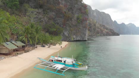 Aerial-drone-shot-over-Paraw-boat-and-towards-a-Philippine-beach,-with-people-swimming-and-enjoying-a-warm-day,-at-Coron-island,-in-Philippines,-Asia