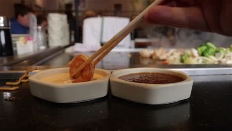 Close-up-of-a-hand-using-chopsticks-to-dip-shrimp-into-sauce-next-to-a-Teppanyaki-grill-at-a-Japanese-restaurant
