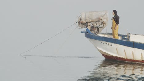 Pequeño-Barco-Pescador-Recogiendo-Redes-De-Pesca-Mientras-Un-Trabajador-Fuma-Un-Cigarrillo-En-El-Mar-Tranquilo-Al-Amanecer-En-Cámara-Lenta-60fps