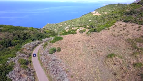 Drone-Aerial-shot,-following-a-car-on-the-coastal-road-on-a-mediterranean-landscape-with-stunning-blue-water-and-green-hills,-in-Sardinia,-Italy-south-of-thes-Spanish-town-of-Alghero