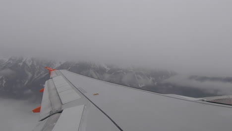 Plane-wing-as-plane-takes-off-flying-over-cloudy-snowy-mountains-in-Austria