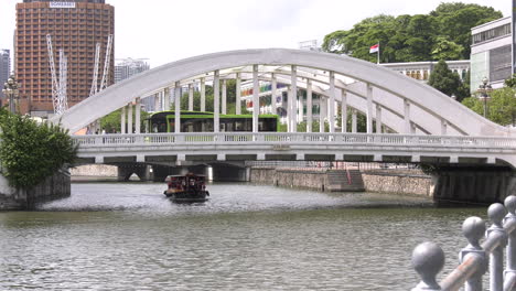 A-boat-ferrying-tourists-going-under-a-bridge-with-cars-driving-by,-Singapore-river,-Boat-Quay