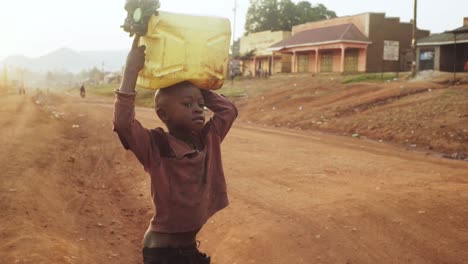 Kleines-Kind-Wartet-Darauf,-Eine-Unbefestigte-Straße-Zu-überqueren,-Während-Es-Bei-Sonnenaufgang-Im-Ländlichen-Uganda-Einen-Wasserkanister-Auf-Seinem-Kopf-Balanciert