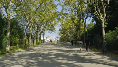 Young-man-standing-and-posing-in-middle-of-park-road-with-people-walking-at-Montjuic-National-Palace,-Palau-Nacional-in-Barcelona,-Spain-on-beautiful-sunny-day-with-trees-and-lanterns-in-park-area