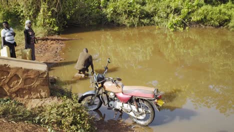 Ein-Mann-Holt-Wasser-Aus-Einem-Verseuchten-Teich,-Während-Zwei-Frauen-In-Der-Schlange-Stehen-Und-Im-Vordergrund-Ein-Fahrrad-Im-Ländlichen-Uganda-Steht
