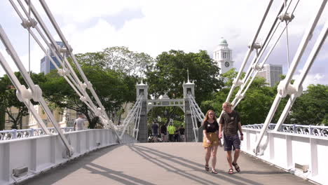 People-walking-across-the-Singapore-river-on-Cavenagh-bridge,-clock-tower-and-trees-in-the-background