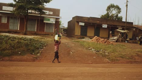 Junges-Ugandisches-Kind,-Das-Mit-Einem-Gelben-Wasserkanister-Auf-Dem-Kopf-Eine-Unbefestigte-Straße-überquert
