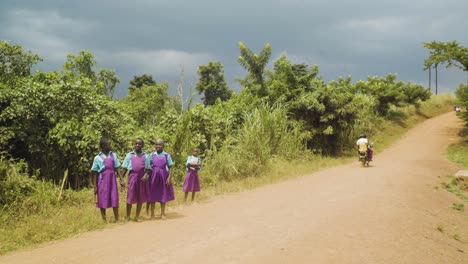 Cuatro-Niñas-De-La-Escuela-De-Pie-Junto-A-Un-Camino-De-Tierra-Con-Bicicletas-Pasando-Cerca-De-Entebbe,-Uganda