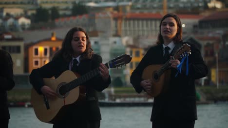 Plano-Medio,-Dos-Mujeres-Jóvenes-Tocando-La-Guitarra-Y-Cantando,-Vista-Panorámica-Del-Edificio-De-Oporto-Y-El-Río-Duero-Al-Fondo
