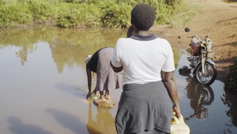 Zwei-Frauen-Holen-Wasser-Aus-Derselben-Pfütze,-In-Der-Ein-Mann-Im-Ländlichen-Uganda-Sein-Fahrrad-Gewaschen-Hat
