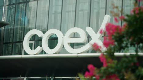 Logo-Des-Coex-Kongress--Und-Ausstellungszentrums-Am-Gebäudeeingang-–-Langsamer-Blick-Hinter-Blühenden-Büschen-–-Kardanische-Aufnahme