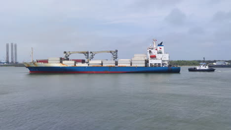 Ein-Riesiges-Containerschiff-Beherrscht-Die-Bucht,-Während-Es-Sich-Elegant-Einem-Industriehafen-Nähert,-Gefolgt-Von-Einem-Speziellen-Schlepper,-Der-Dicht-Dahinter-Folgt,-Mit-Malerischer-Aussicht,-Industriehafen,-Logistik,-Containerschiff