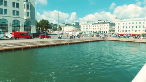 Bootsfahrt-Von-Hinten-Im-Hafen-Von-Helsinki,-Abfahrt-Mit-Stadtgebäuden-Und-Menschen,-Die-Am-Pier-Entlanglaufen
