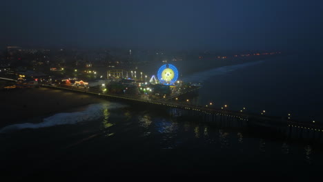 Drone-shot-approaching-the-glowing-Ferris-wheel-on-Santa-Monica-pier,-blue-hour-in-LA,-USA