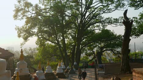 Landscape-view-of-people-walking-in-a-hindu-temple-area-with-many-trees,-in-Kathmandu,-Nepal,-at-sunset