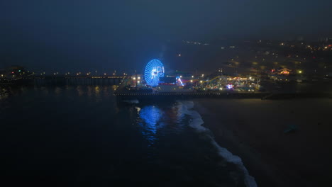Aerial-view-away-from-the-illuminated-Santa-Monica-pier,-low-visibility-evening-in-LA,-USA---pull-back,-drone-shot